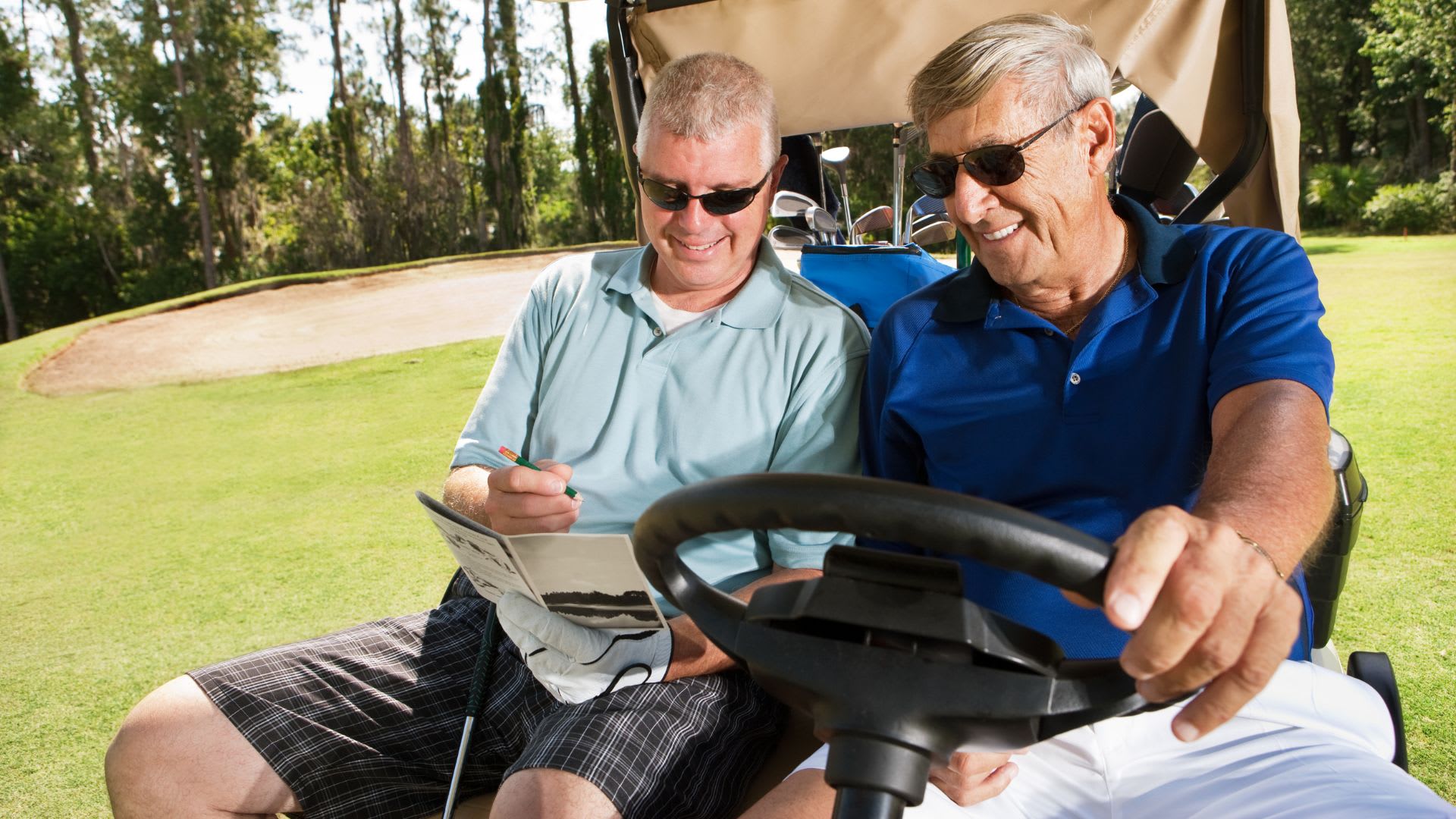 Two men sit in a golf cart, ready to tally their scores. One, in a light blue shirt, holds a pen and paper for tracking birdies and bogeys, while the other drives in his dark blue shirt. Both sport sunglasses as they prepare for the next hole.